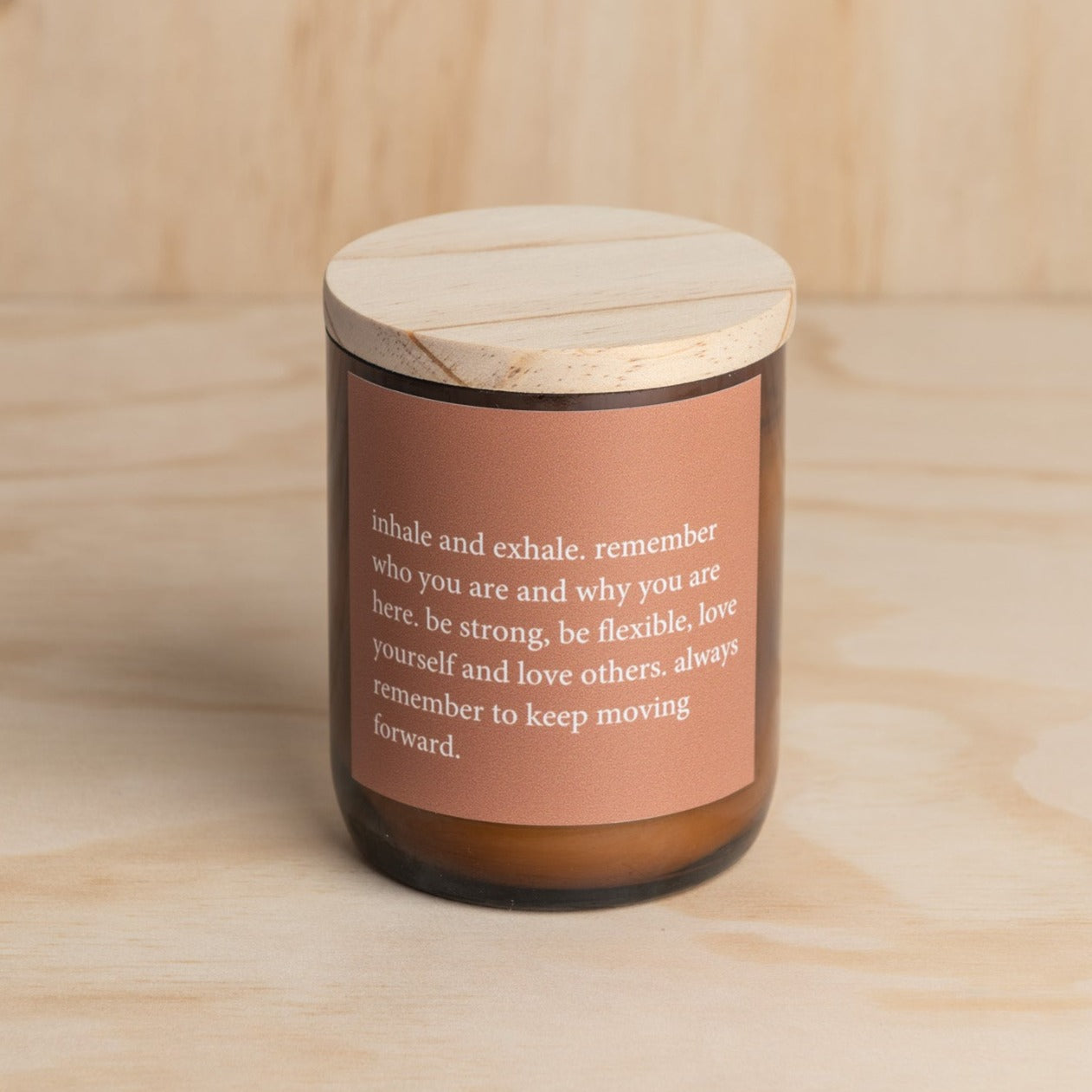 The Commonfolk scented candle has a brown jar and a wooden lid, featuring a quote around the middle in a white font.