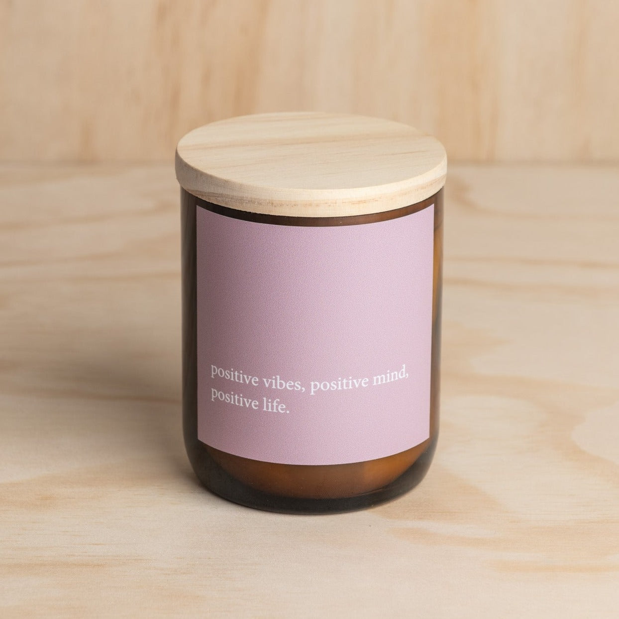 The Commonfolk scented candle has a purple label that reads 'positive vibes, positive mind, positive life.' The candle is placed in an amber jar with a wooden lid.