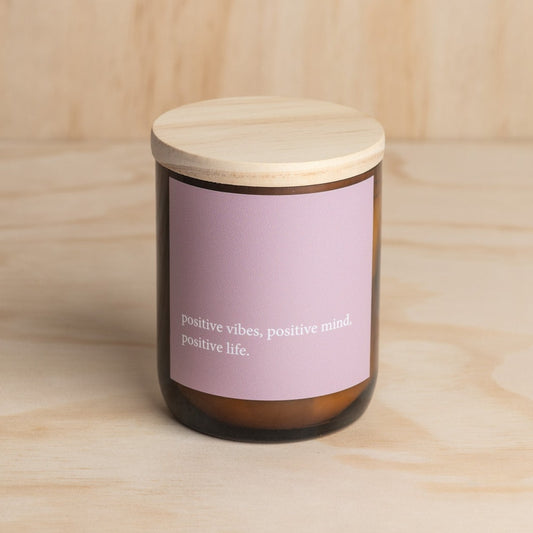 The Commonfolk scented candle has a purple label that reads 'positive vibes, positive mind, positive life.' The candle is placed in an amber jar with a wooden lid.