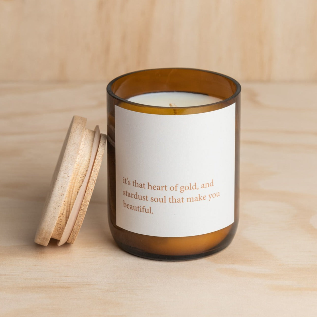 The Commonfolk scented soy wax candle is in an amber glass jar with a white label, featuring the quote 'it's that heart of gold, and stardust soul that make you beautiful'. The lid is placed next to the jar.