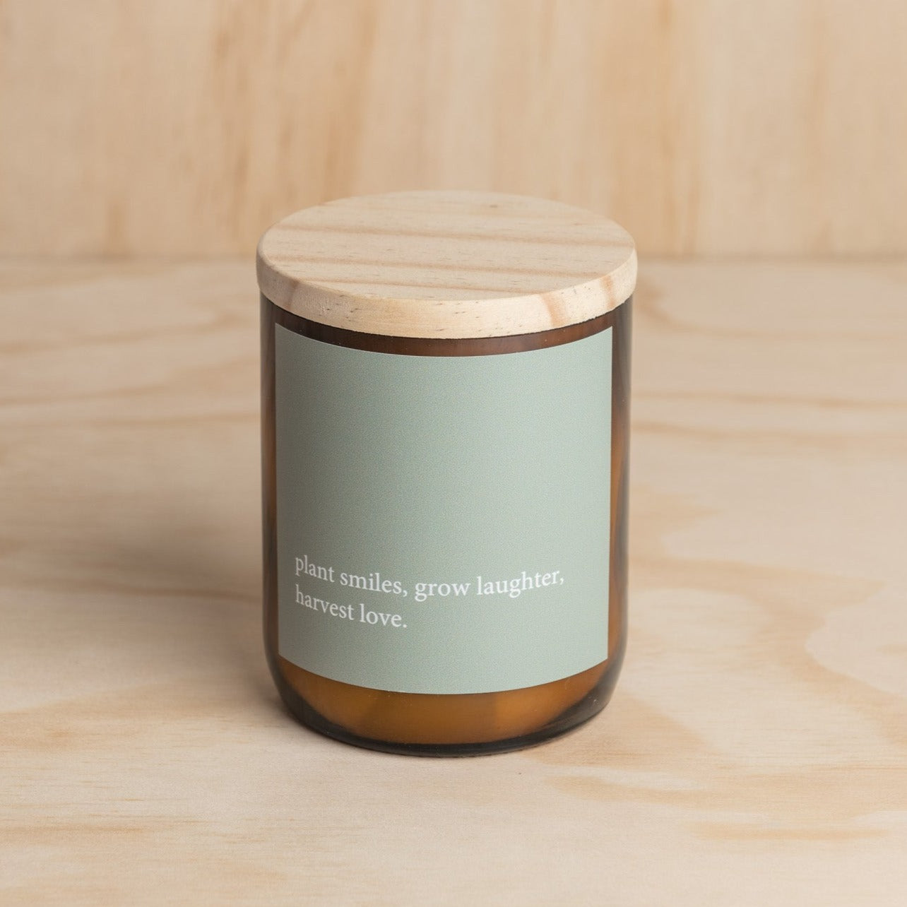 The Commonfolk scented mid-sized amber jar candle with a wooden lid, features a green label with the quote 'plant smiles, grow laughter, harvest love.'.