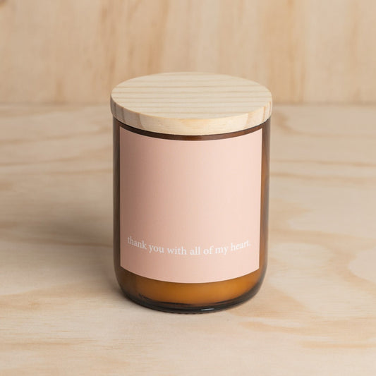 The Commonfolk scented midsize amber jar candle with a wooden lid and a label that reads 'thank you with all of my heart'. The candle is set against a wooden background.