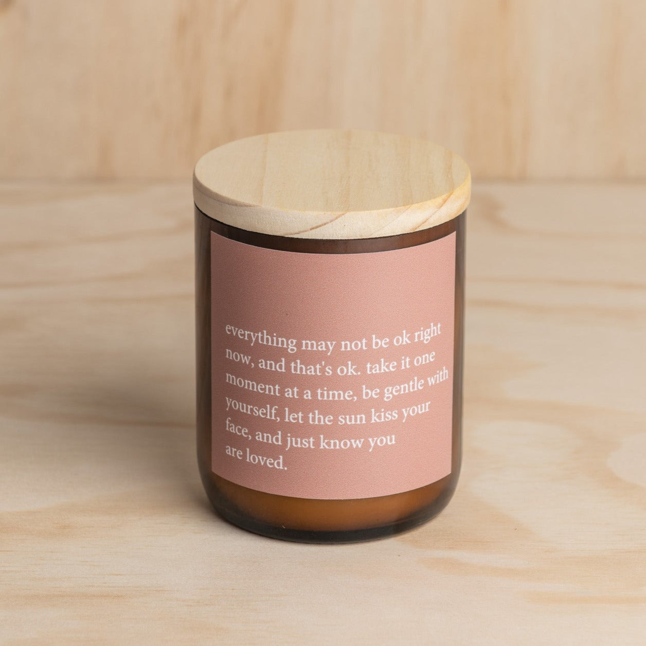 The Commonfolk scented soy candle in an amber jar with a wooden lid features a quote on the pink label.