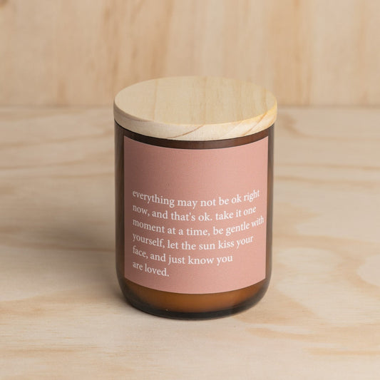 The Commonfolk scented soy candle in an amber jar with a wooden lid features a quote on the pink label.