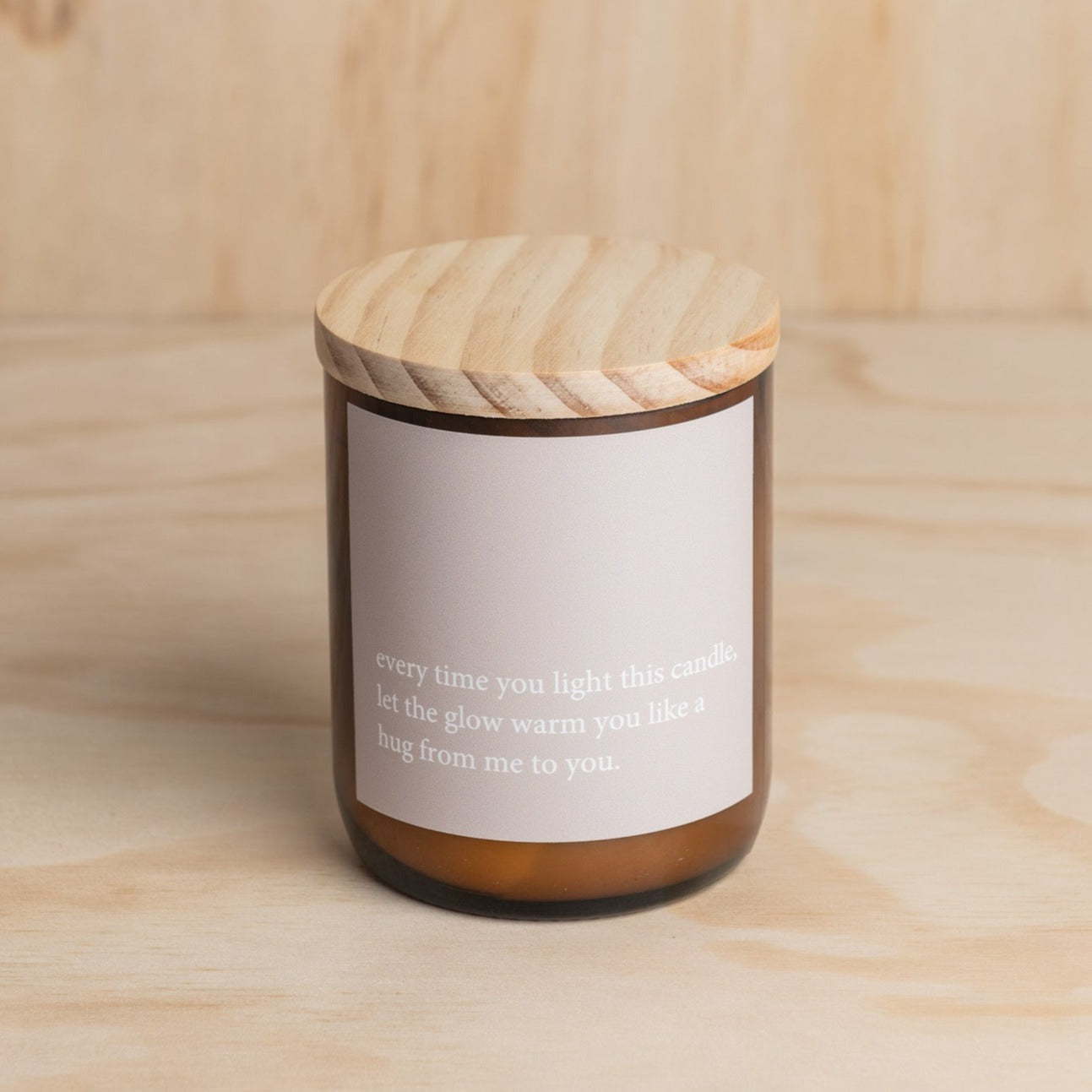 The Commonfolk scented mid-sized amber jar candle with a wooden lid features a white label with a heartfelt quote.
