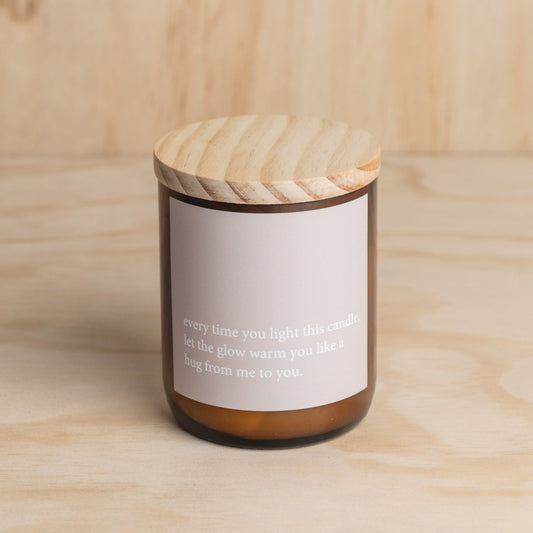 The Commonfolk scented mid-sized amber jar candle with a wooden lid features a white label with a heartfelt quote.