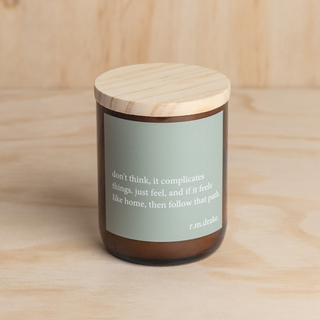 The Commonfolk scented candle with a wooden lid and a green label featuring a heartfelt quote is placed on a wooden surface.