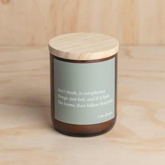 The Commonfolk scented candle with a wooden lid and a green label featuring a heartfelt quote is placed on a wooden surface.
