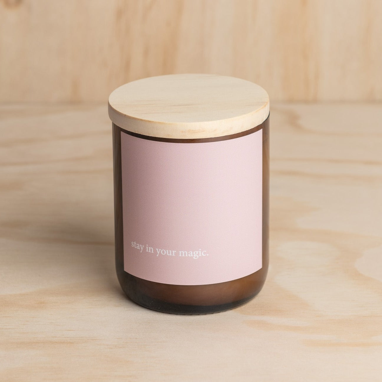 The Commonfolk scented candle has a wooden lid and a label that reads 'stay in your magic'. The body of the candle is a dark color, likely brown, and the label is in a light color, likely pink or beige.