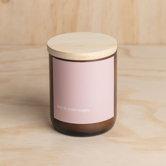 The Commonfolk scented candle has a wooden lid and a label that reads 'stay in your magic'. The body of the candle is a dark color, likely brown, and the label is in a light color, likely pink or beige.