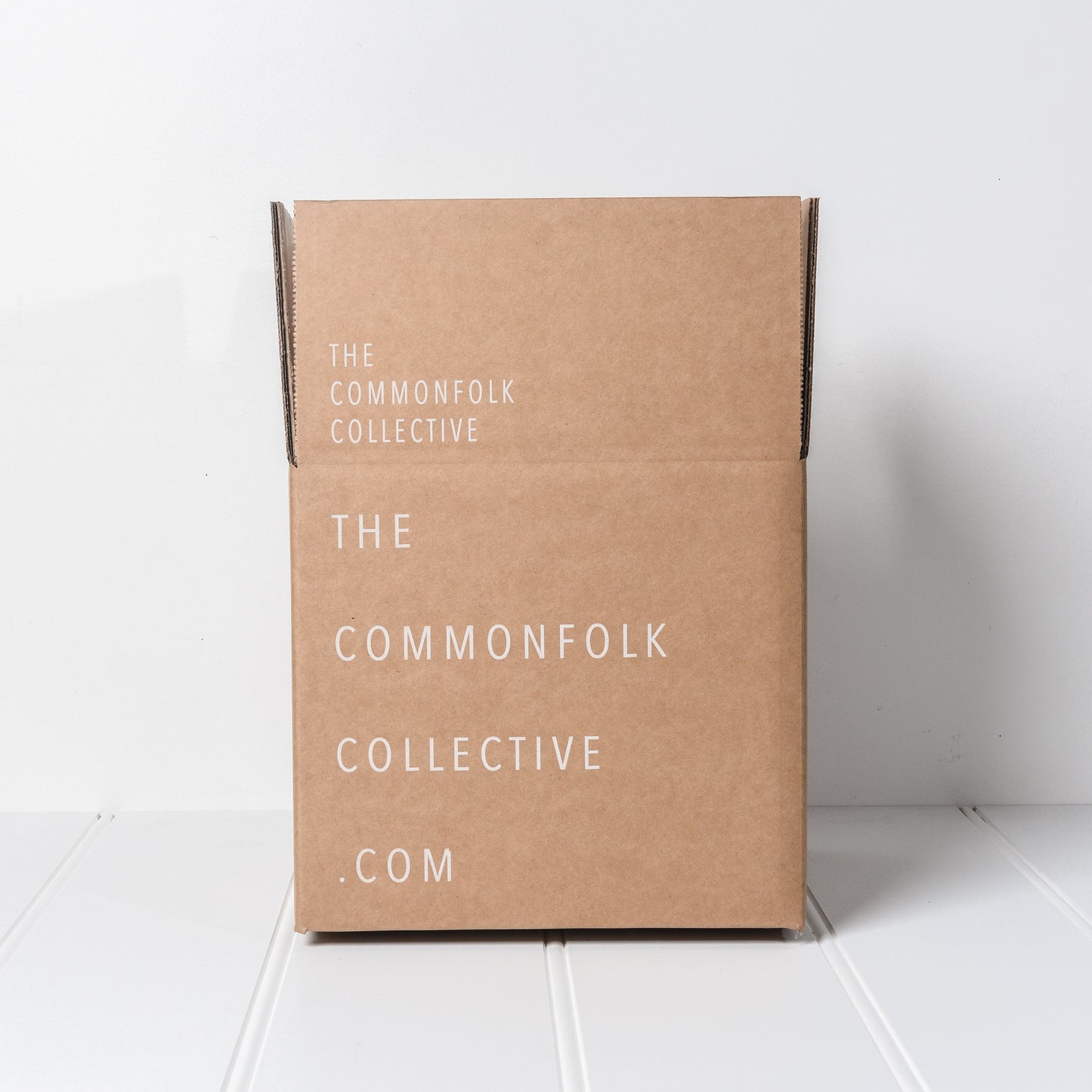 A brown cardboard gift box with 'THE COMMONFOLK COLLECTIVE.COM' printed on the side, indicating it is from the Commonfolk Collective brand.