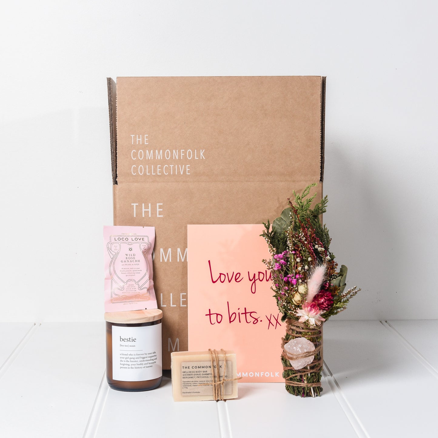 The Commonfolk gift box contains a midi quote candle, a dried flower posy with a rose quartz crystal, an Australian-made wrapped body bar/soap, Loco Love chocolate, and a card with a personalized message.