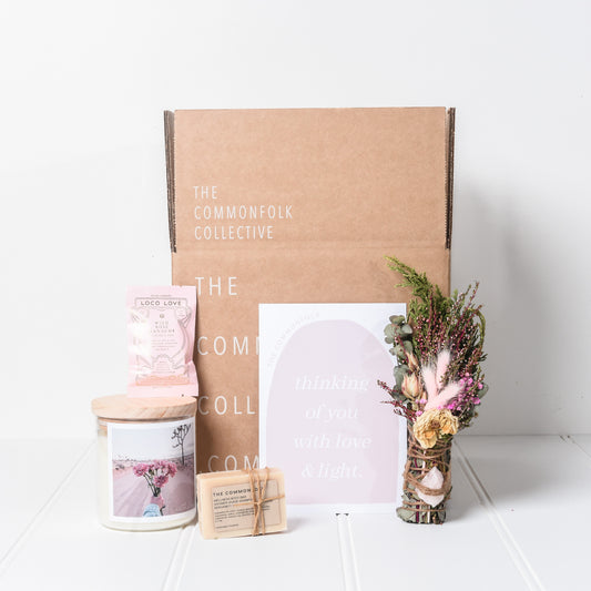 The Commonfolk deluxe gift box set includes a large candle, a dried flower posy with a rose quartz crystal, a wrapped body bar soap, a chocolate bar, and a giant gift card with a personal message, all displayed next to a cardboard box.