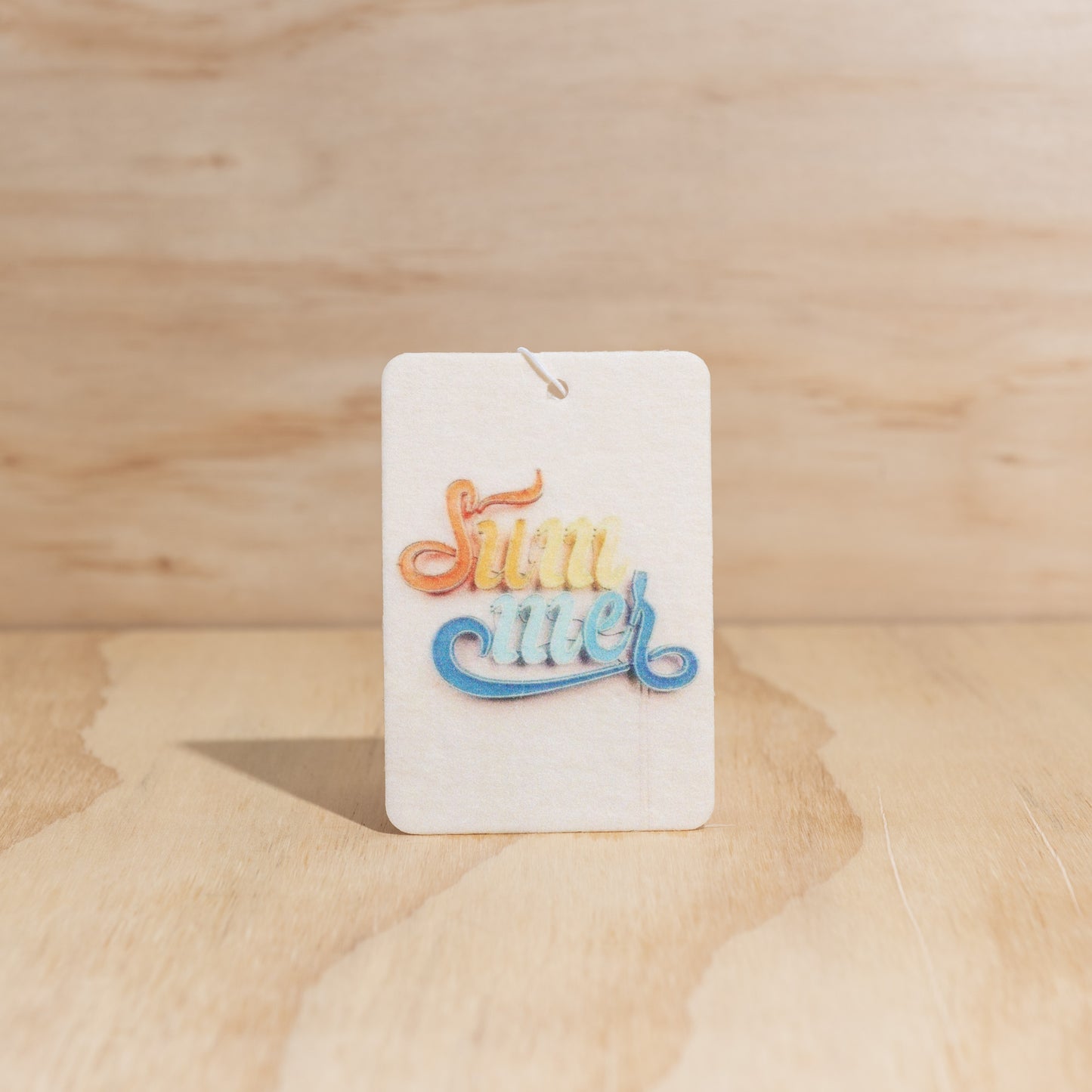 The Commonfolk air freshener with the text 'SUMMER' in bold, multicolored letters on a white background, displayed against a wooden backdrop.