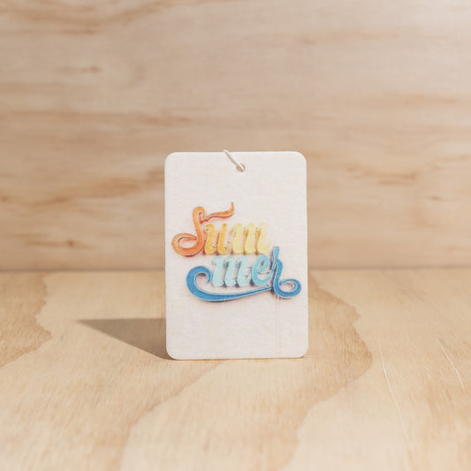 The Commonfolk air freshener with the text 'SUMMER' in bold, multicolored letters on a white background, displayed against a wooden backdrop.