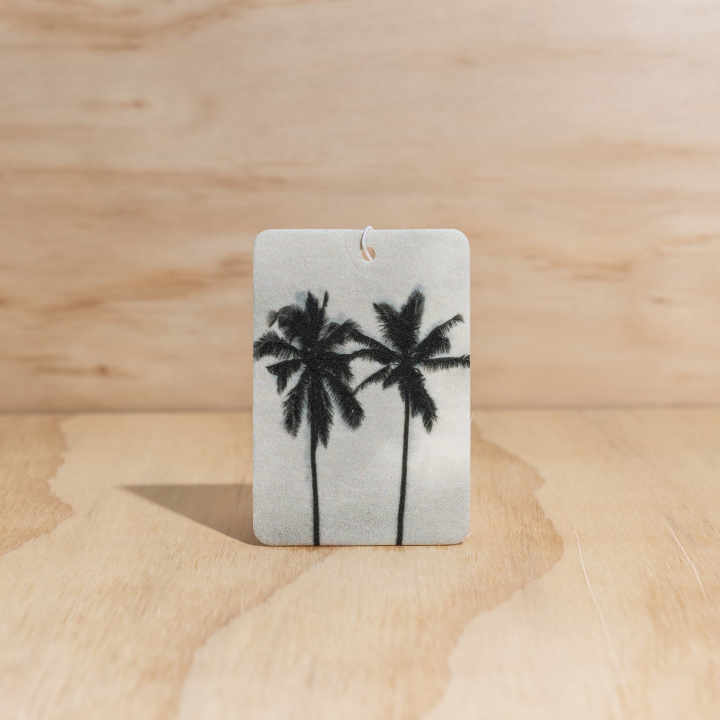 The Commonfolk air freshener, with a white background featuring black silhouettes of palm trees, displays on a wooden background.