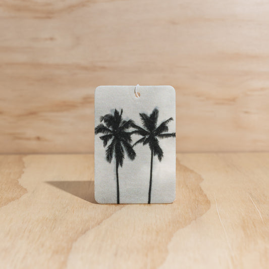 The Commonfolk air freshener, with a white background featuring black silhouettes of palm trees, displays on a wooden background.