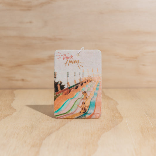 The Commonfolk air freshener with a colorful design and text reading 'Think Happy', is displayed on a wooden background.