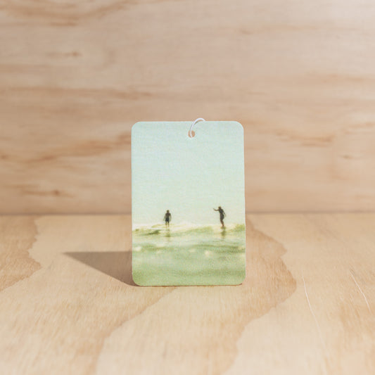 The Commonfolk air freshener, with a depiction of a beach scene, is displayed against a wooden background.