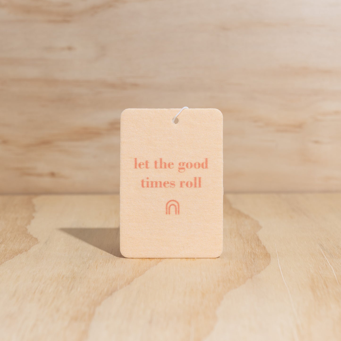 The Commonfolk air freshener with the text 'let the good times roll' printed in orange ink on a beige background, is displayed against a wooden backdrop.