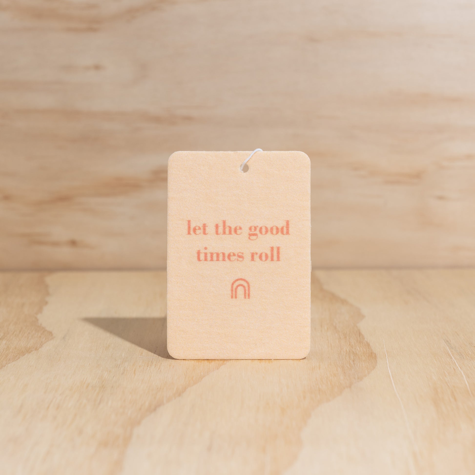The Commonfolk air freshener with the text 'let the good times roll' printed in orange ink on a beige background, is displayed against a wooden backdrop.