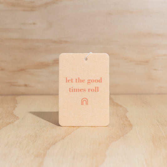 The Commonfolk air freshener with the text 'let the good times roll' printed in orange ink on a beige background, is displayed against a wooden backdrop.