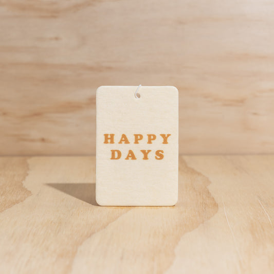 The Commonfolk air freshener with the text 'HAPPY DAYS' printed in brown. It is placed against a wooden background.