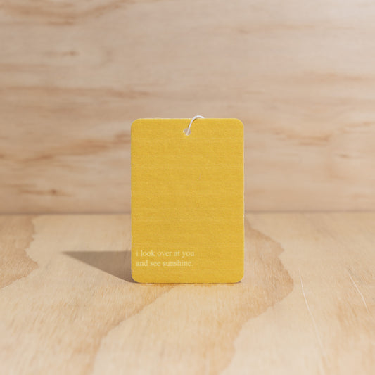 The Commonfolk air freshener features a yellow label with the text 'I look over at you and see sunshine,' displayed against a wooden background.