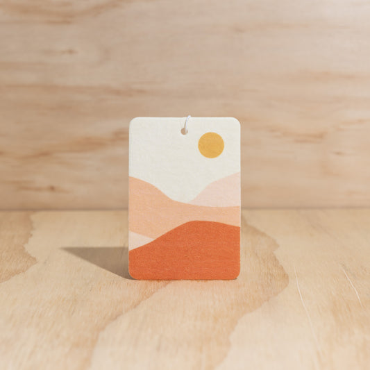 The Commonfolk air freshener with a geometric design featuring a sun and mountains in shades of white, orange, and brown, displays against a wooden background.
