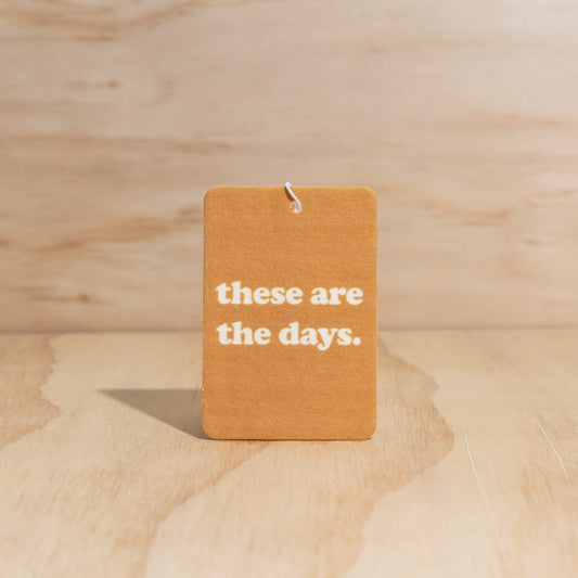 The Commonfolk air freshener with the text 'these are the days.' printed in the center, displays on a wooden surface.