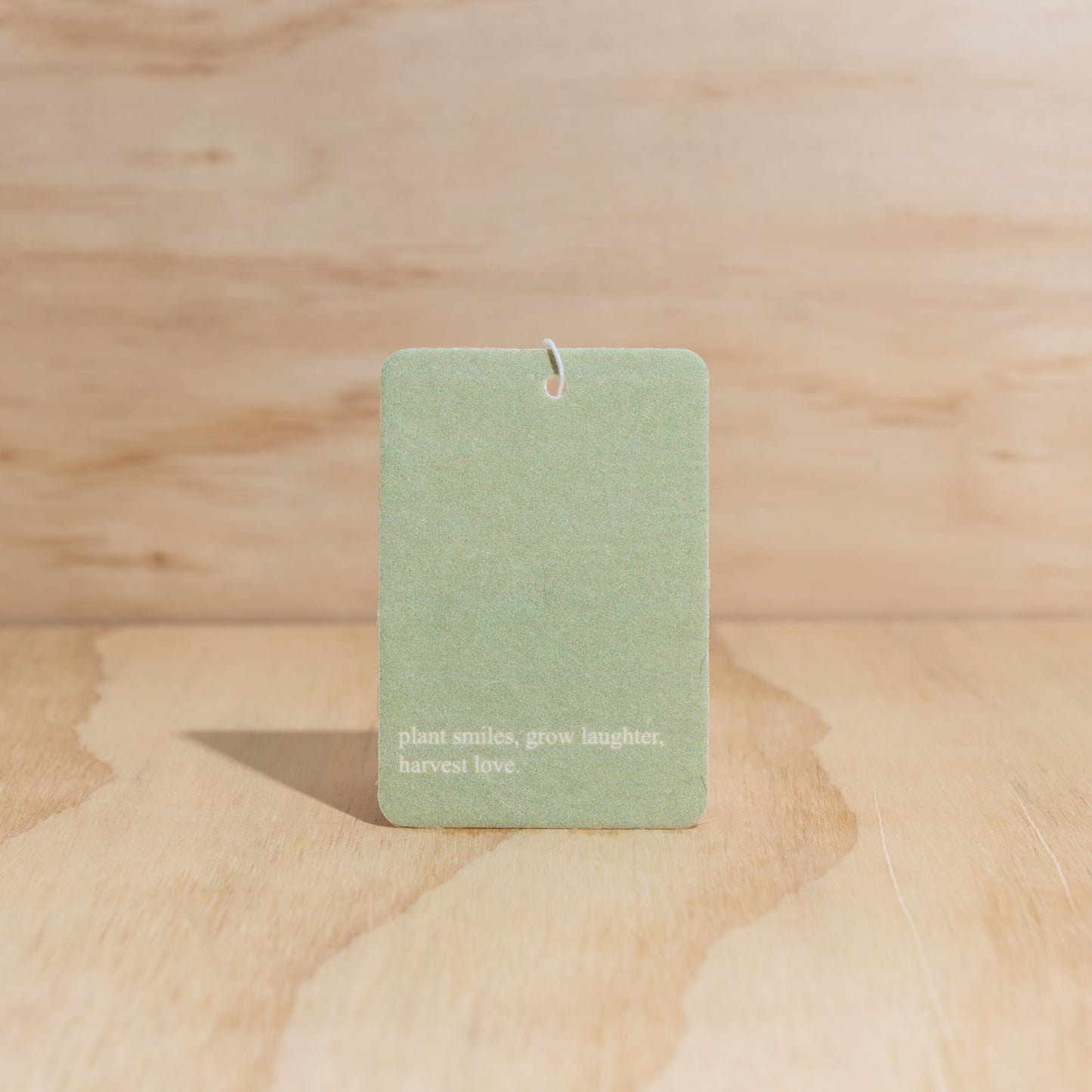 The Commonfolk air freshener features a green label with white text that reads 'plant smiles, grow laughter, harvest love' displayed on a wooden surface.