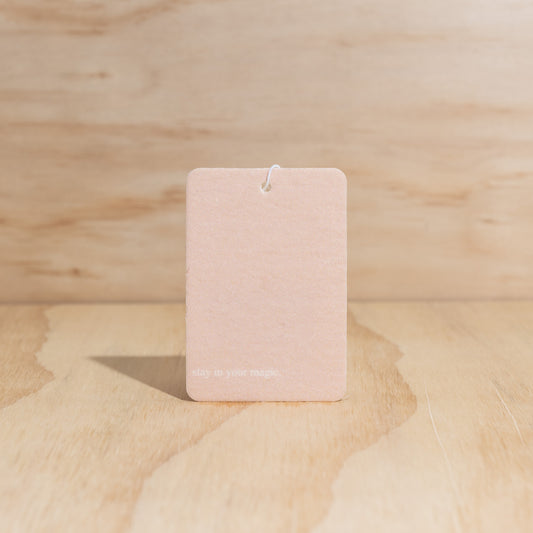 The Commonfolk air freshener features a pink label with a message that reads 'Stay in Your Magic' displayed on a wooden surface.