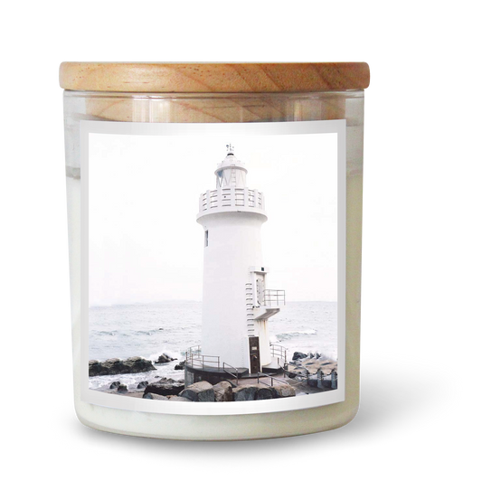 The Commonfolk scented white candle with a lighthouse design is placed in a clear glass jar with a wooden lid.