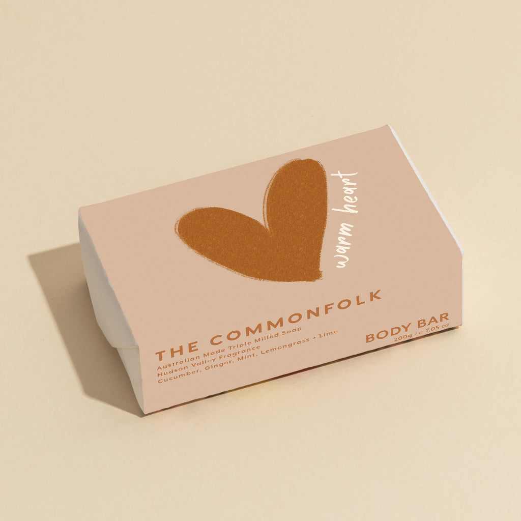 The Commonfolk 200g body bar soap, with a brown heart shape on the top, has packaging in a pinkish hue with the text 'warm heart.'