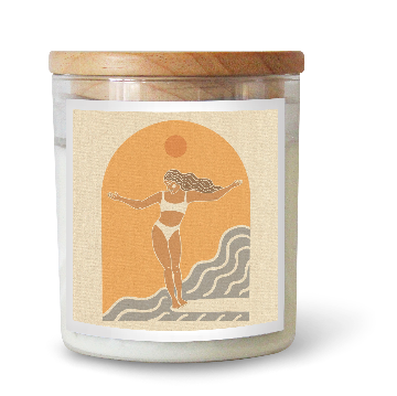 The Commonfolk scented white candle features a label with an illustration of a person standing on a surfboard with outstretched arms, set against a stylized sun and wave background. The candle has a wooden lid.