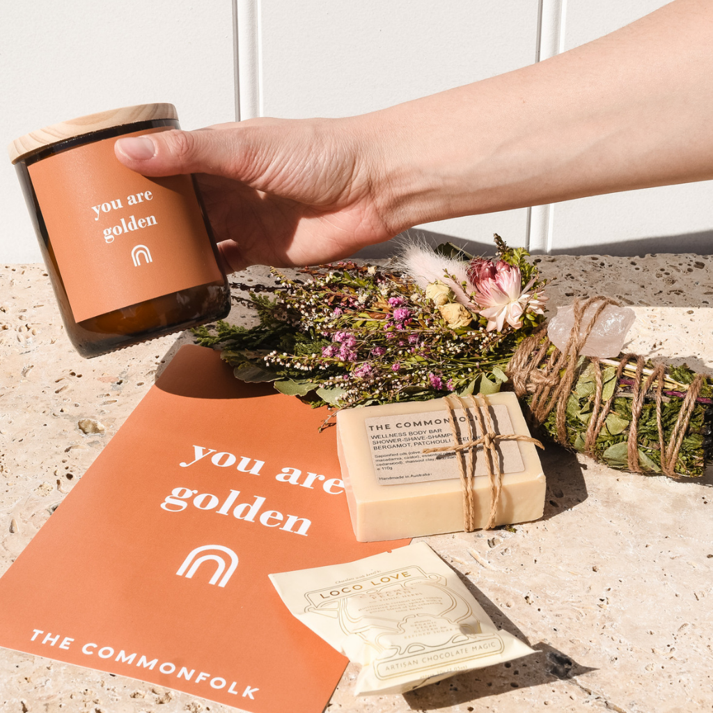A hand holding the scented candle from The Commonfolk, decorative flowers, and a gift card.