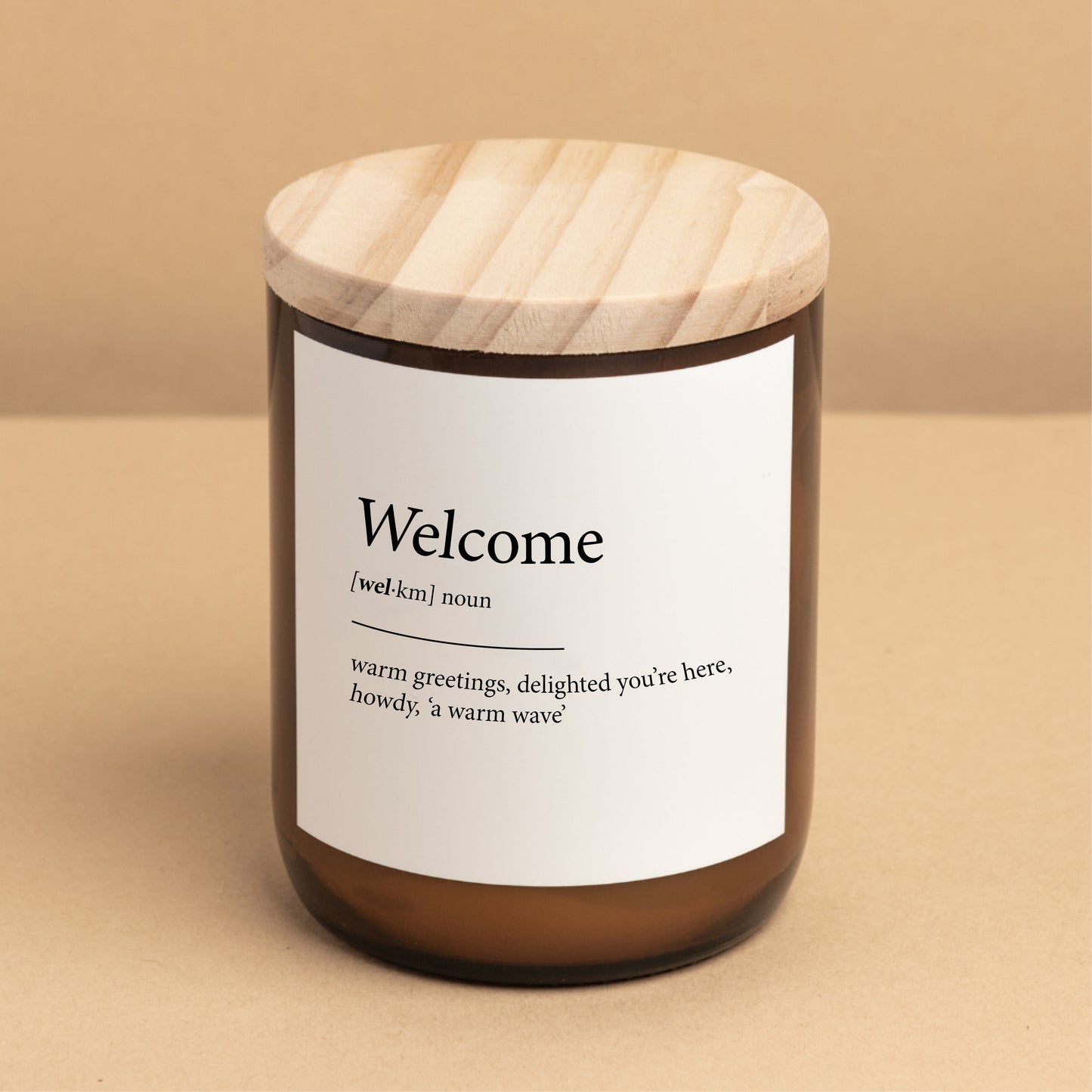 The Commonfolk scented candle, with a label that reads 'Welcome' and showcases its definition, is placed on a minimalist background for a warm ambiance.