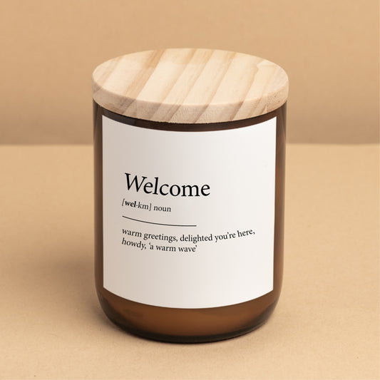 The Commonfolk scented candle, with a label that reads 'Welcome' and showcases its definition, is placed on a minimalist background for a warm ambiance.