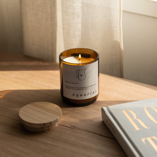 The Commonfolk scented candle with an amber glass jar has a label 'Aquarius' and is placed on a wooden surface, with a wooden lid beside it.