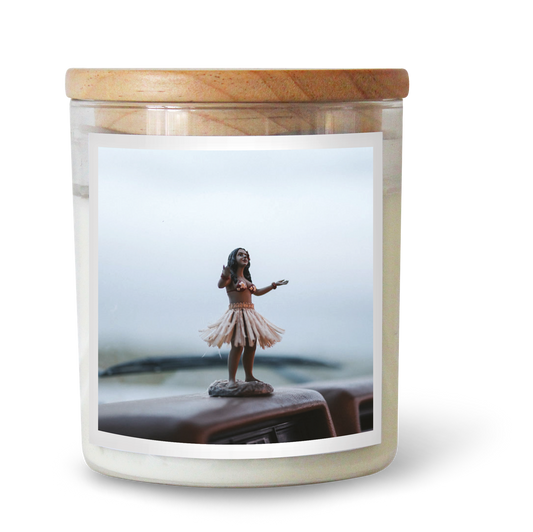 The Commonfolk scented candle with a hula girl figurine on the top is set in a clear jar with a wooden lid.