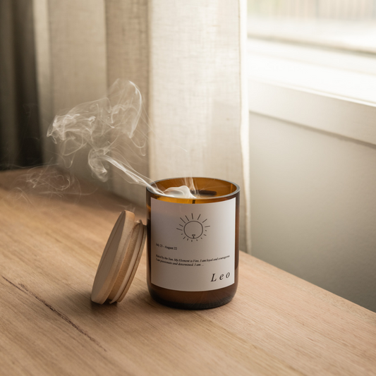 The Commonfolk scented soy wax candle with a lid, labeled 'Leo' and featuring a sun illustration, sitting on a wooden surface with smoke rising from it, indicating it is currently burning or has been burned.