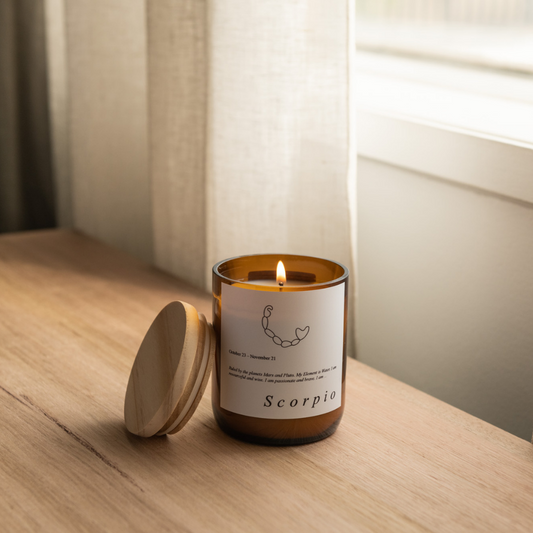 The Commonfolk scented Scorpio-themed candle, with a lit flame, is placed on a wooden surface, featuring a label and a zodiac sign illustration. It comes in an amber glass jar with a wooden lid.