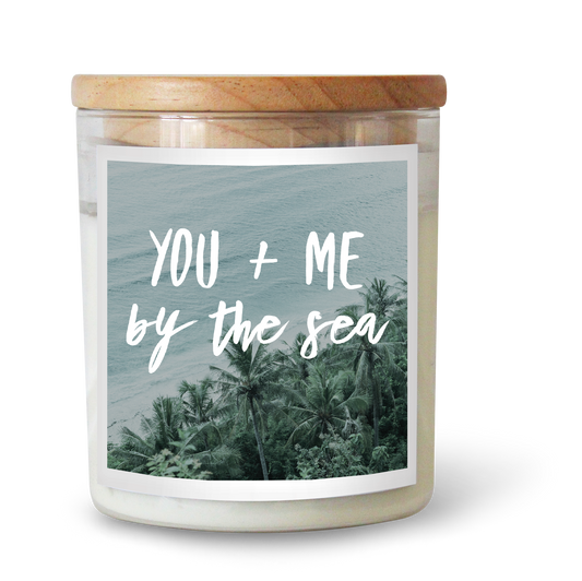The Commonfolk scented white candle with a wooden lid, labeled 'YOU + ME by the Sea' against a background of a tropical beach scene.