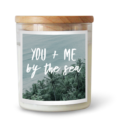 The Commonfolk scented white candle with a wooden lid, labeled 'YOU + ME by the Sea' against a background of a tropical beach scene.