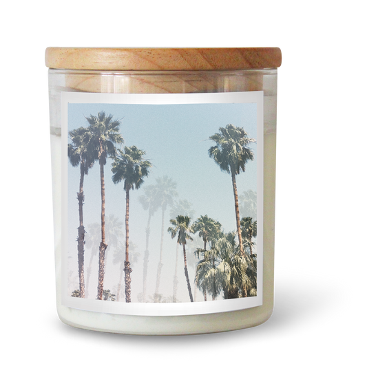 The Commonfolk scented candle is in a clear glass jar with a label featuring a photograph of palm trees and a wooden lid on top.