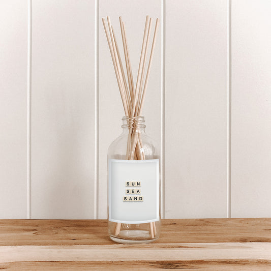The Commonfolk room diffuser with reeds labeled 'Sun Sea Sand' on a wooden surface with a white paneled background