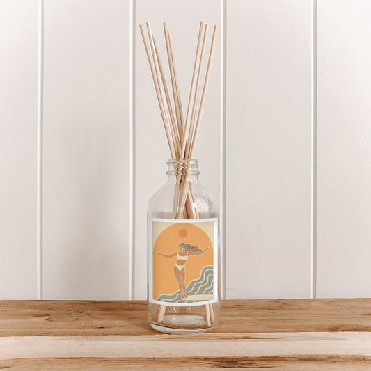 The Commonfolk room diffuser in a clear glass bottle with reeds is on a wooden surface with a light wooden panel background.