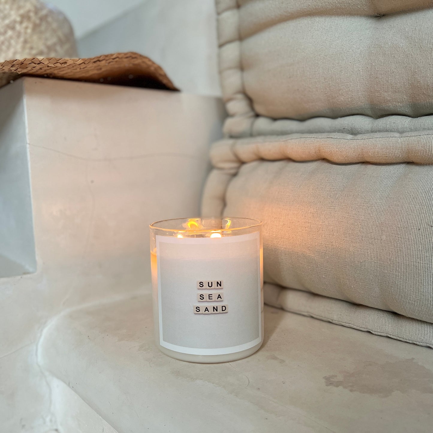 Candle labeled 'Sun Sea Sand' on a stone surface with neutral-toned pillows and a hat in the background.