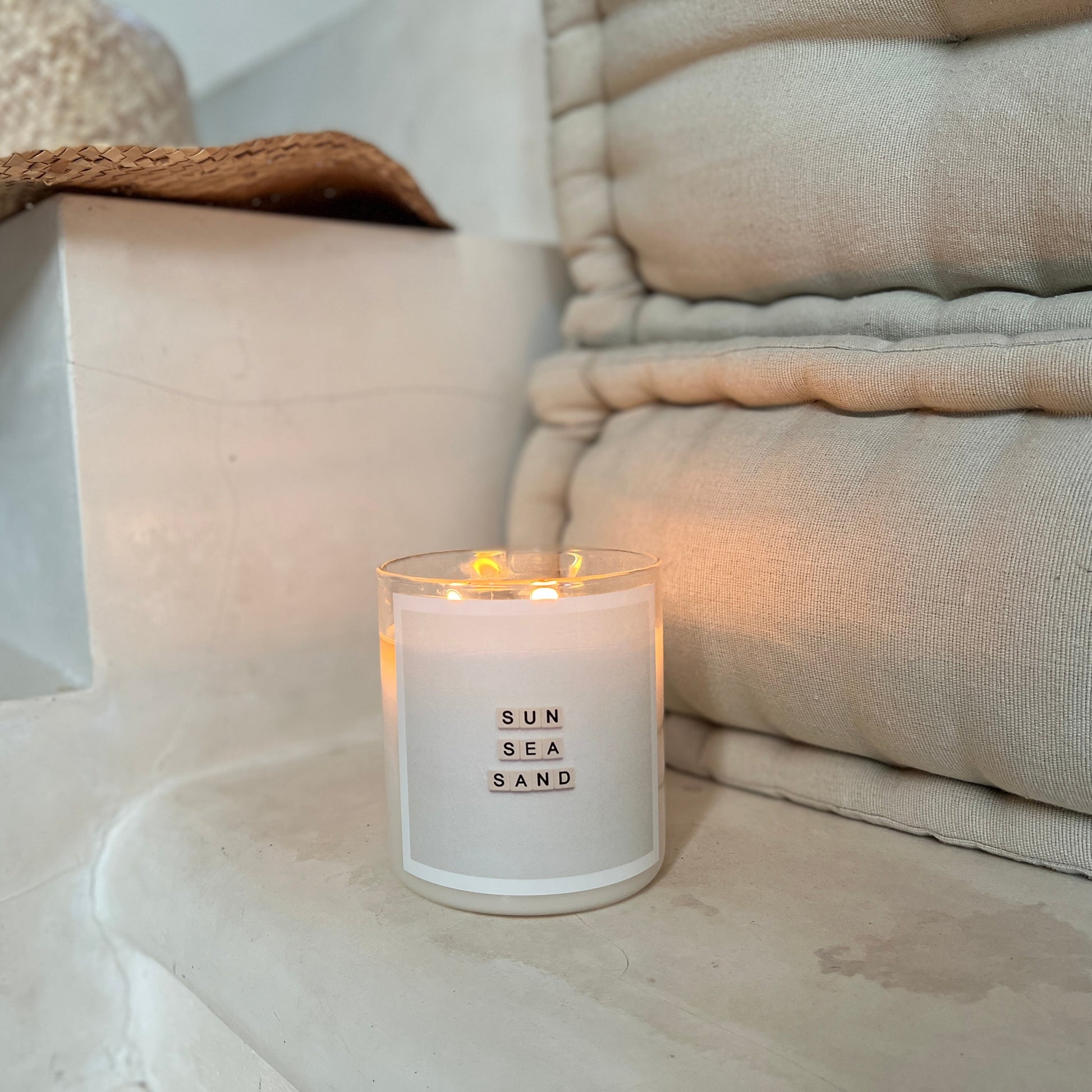 Candle labeled 'Sun Sea Sand' on a stone surface with neutral-toned pillows and a hat in the background.