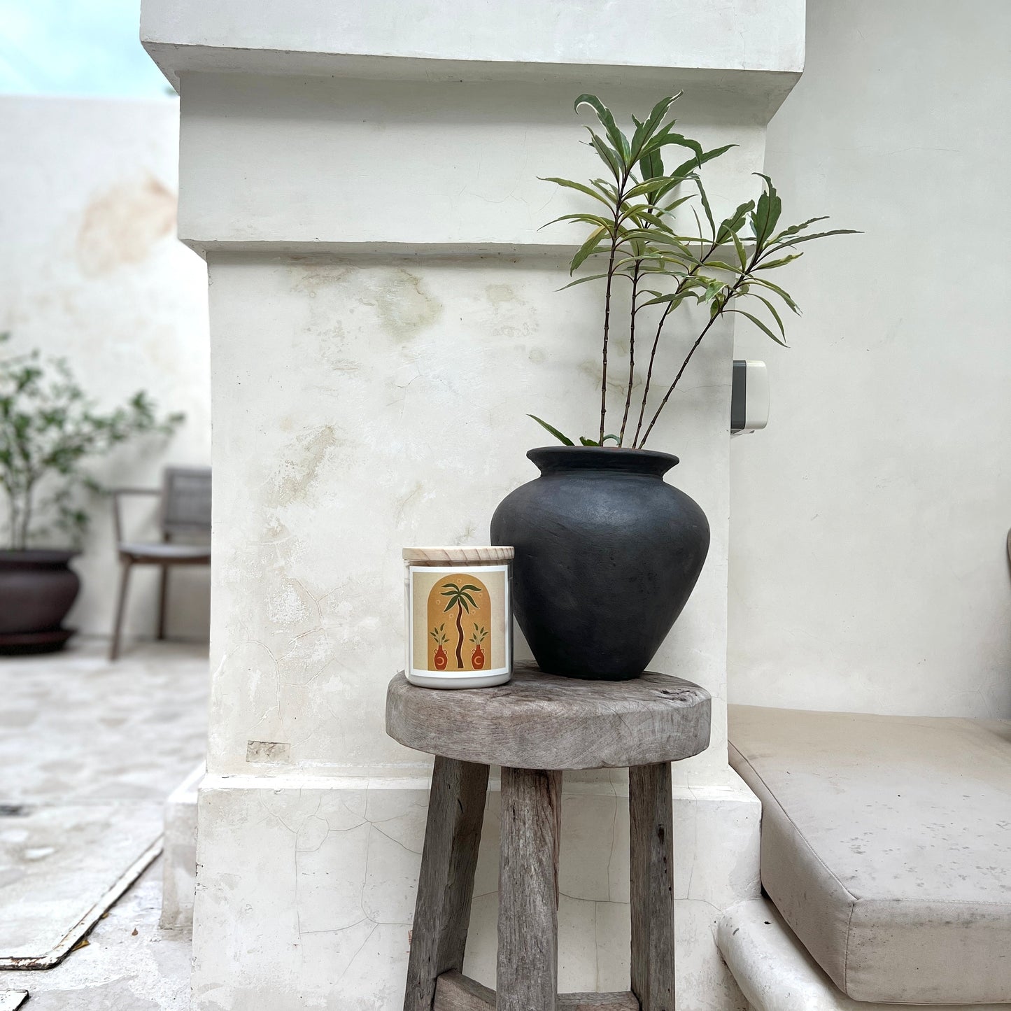 Candle with Black vase with a plant on a wooden stool against a white wall.